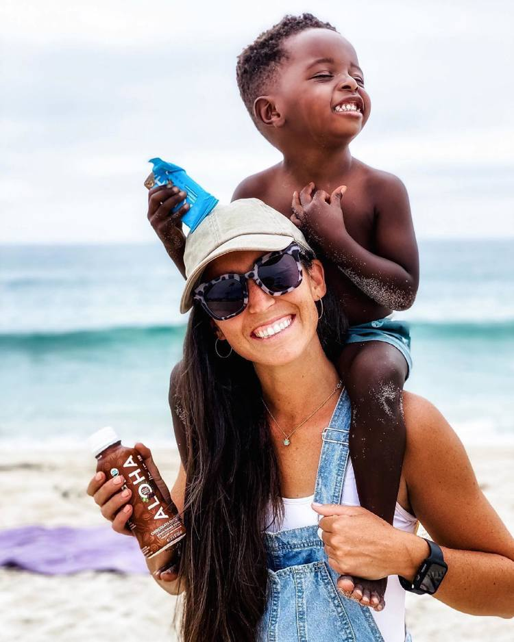 a woman is carrying a child on her shoulders on the beach while holding a bottle of aloha