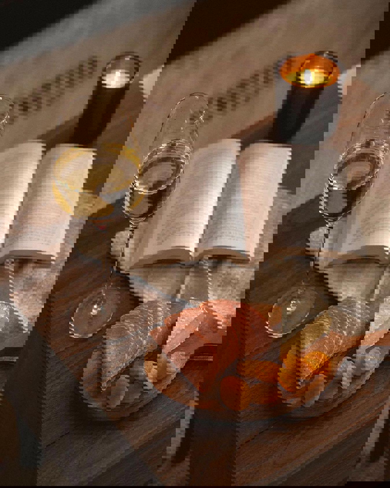 a glass of wine , a book , a cutting board with meat and cheese , and a candle on a table