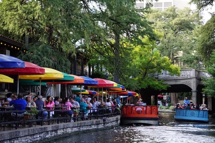 a group of people are sitting at tables under colorful umbrellas next to a river