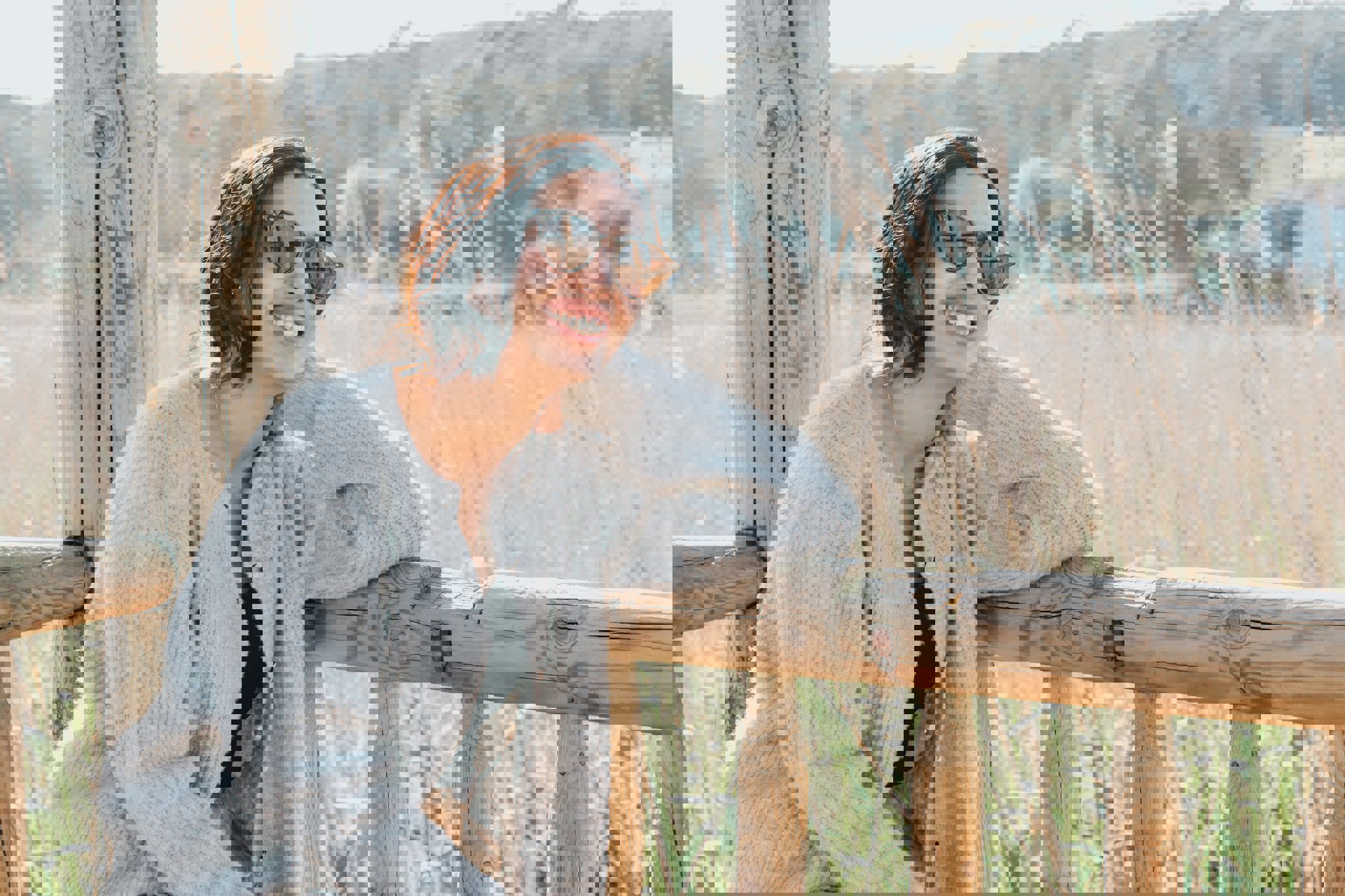 a woman wearing sunglasses and a grey sweater smiles while leaning on a wooden railing