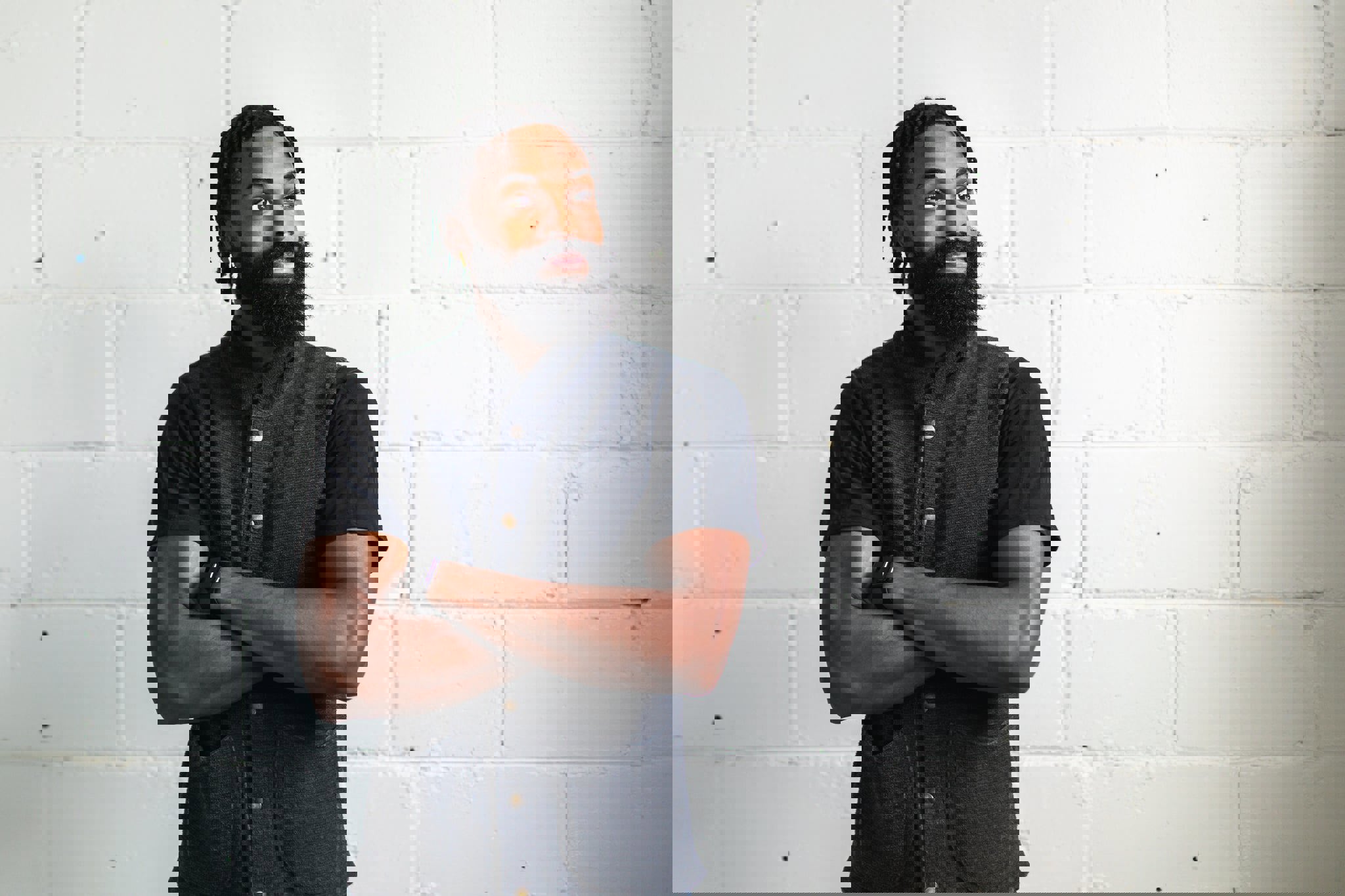 a man with a beard is standing in front of a white brick wall with his arms crossed